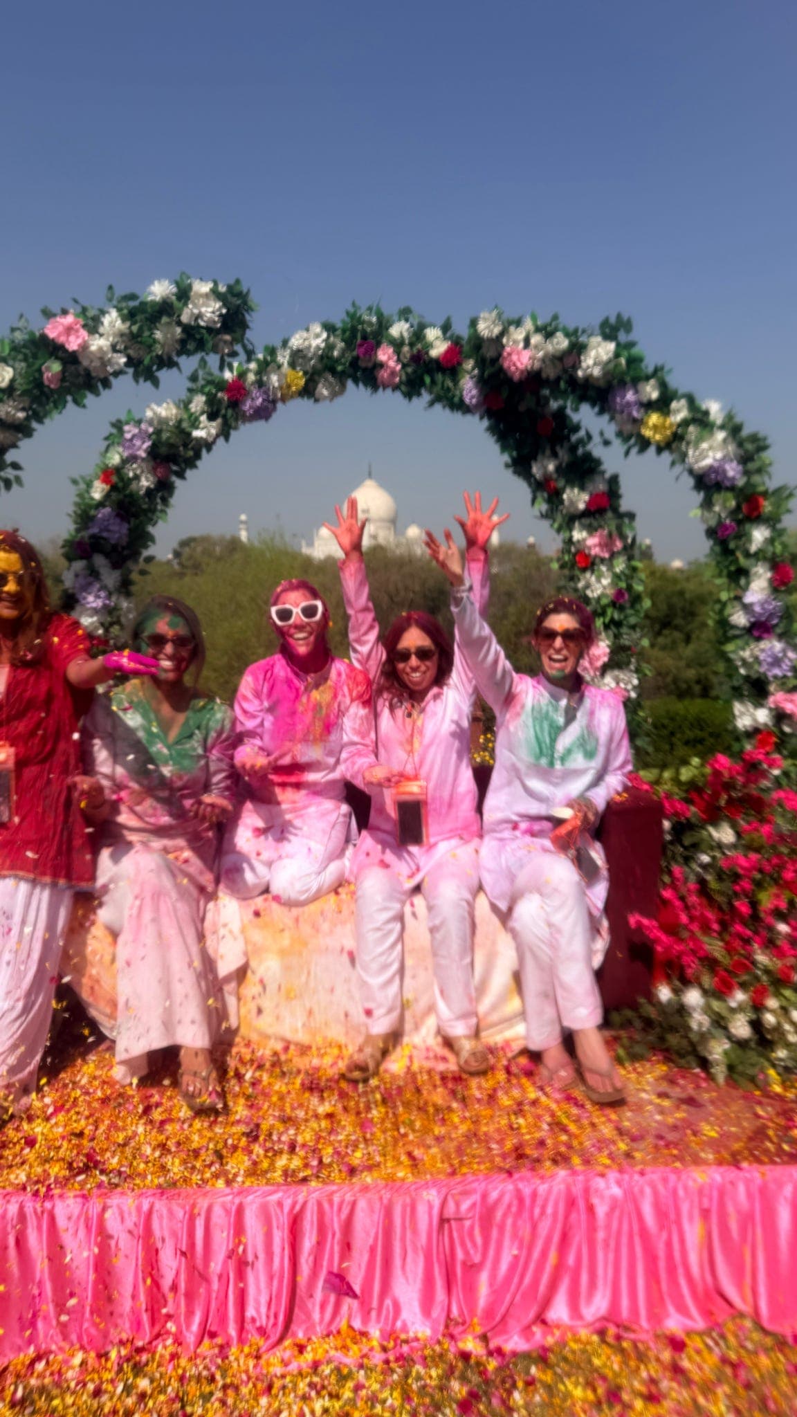 Travellers raising colour-covered hands beneath a floral arch framing the Taj Mahal