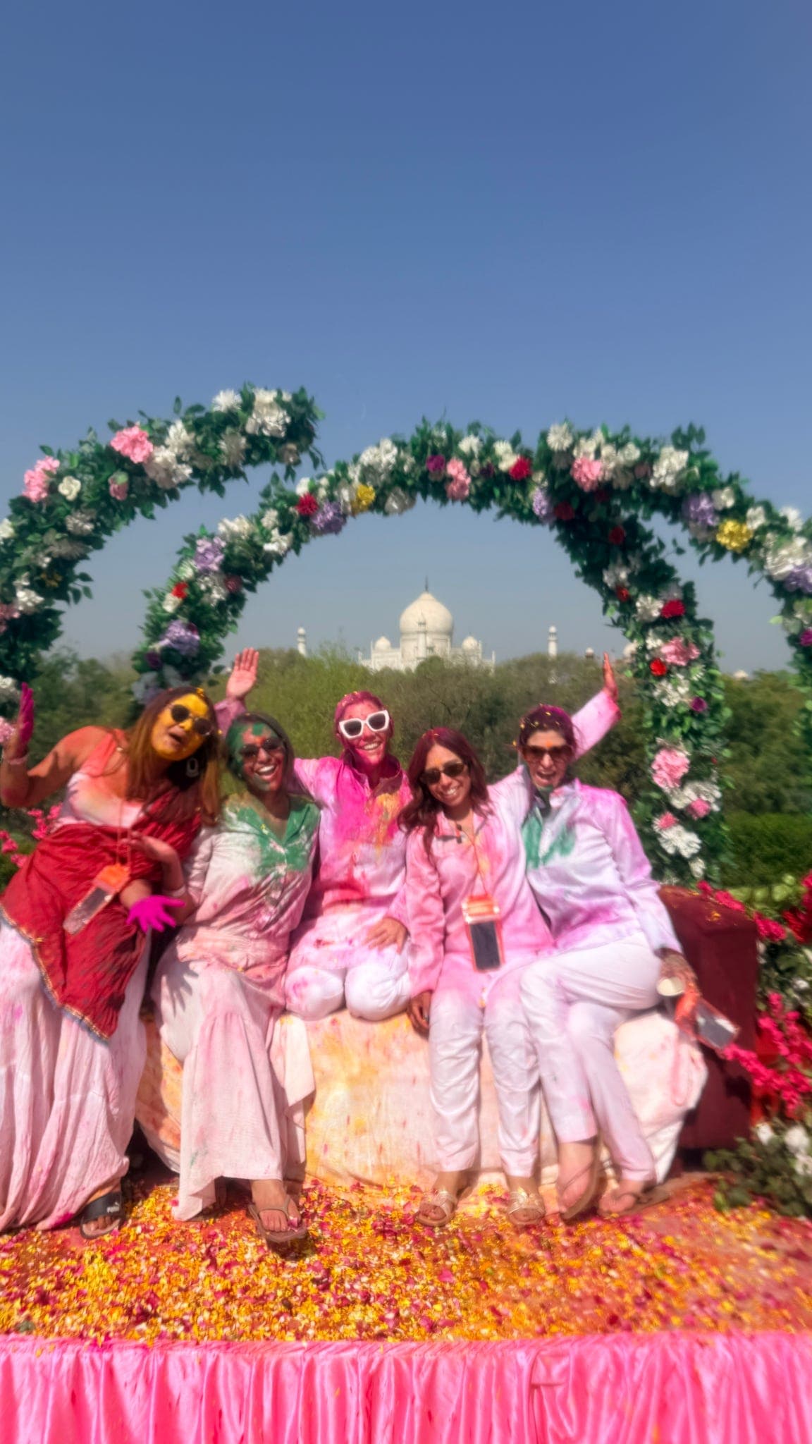 The small group waving in Holi colours on a petal-strewn stage with Taj Mahal views
