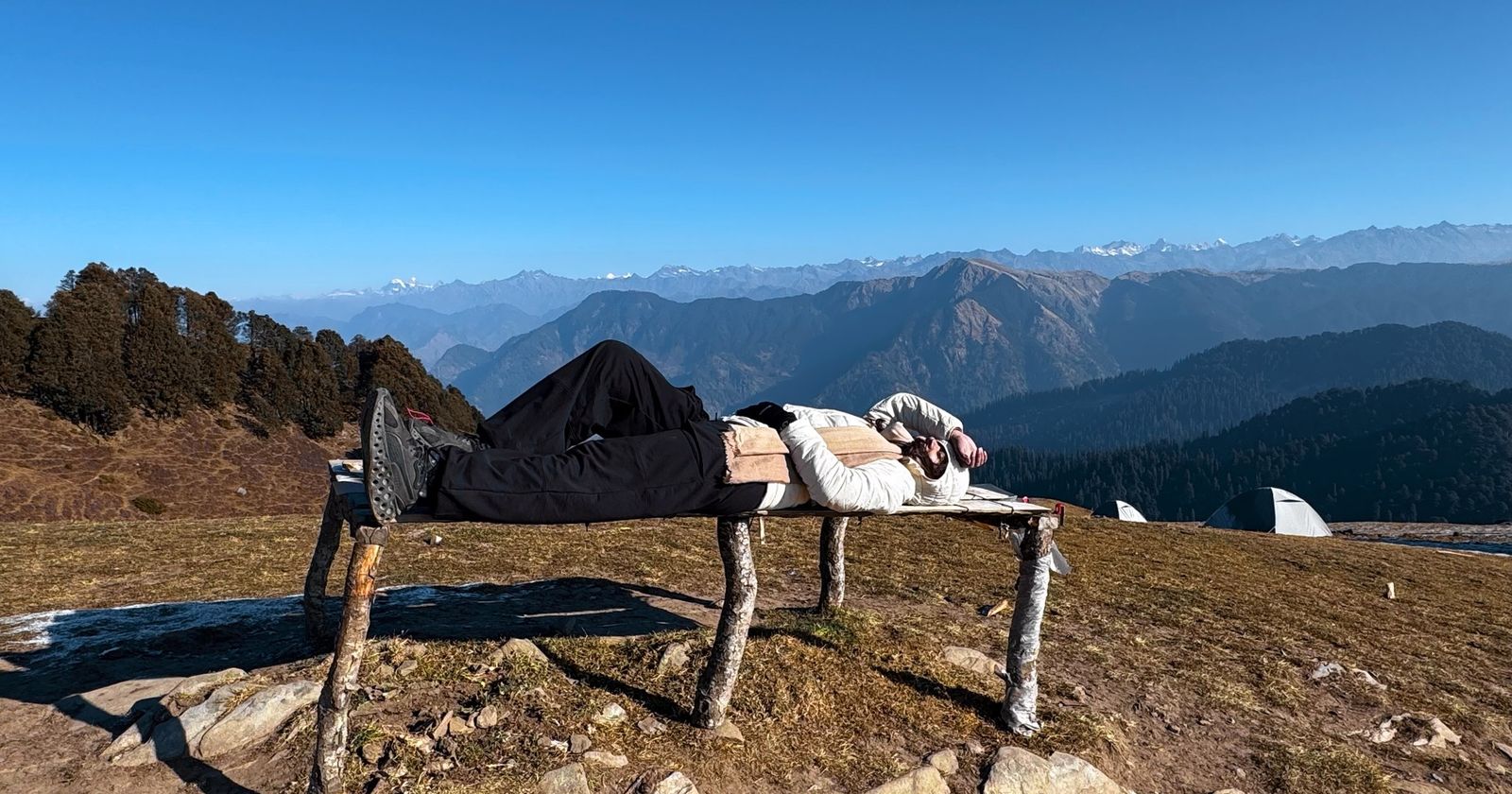 A traveller resting on a wooden charpai on a high meadow with the Himalayan range behind