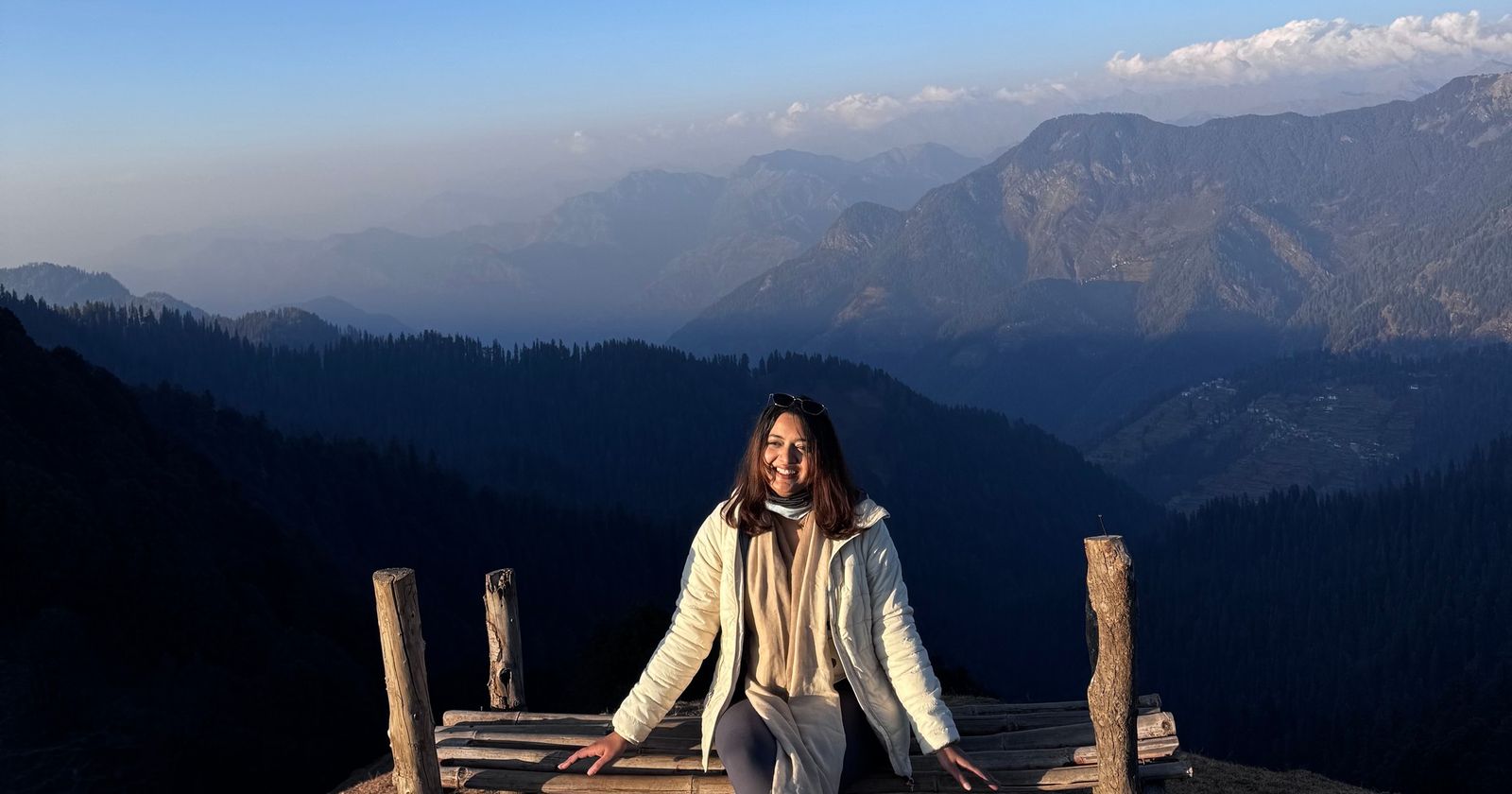 A woman sitting on a wooden bench at the edge of a Himalayan ridge, with layered mountains behind her
