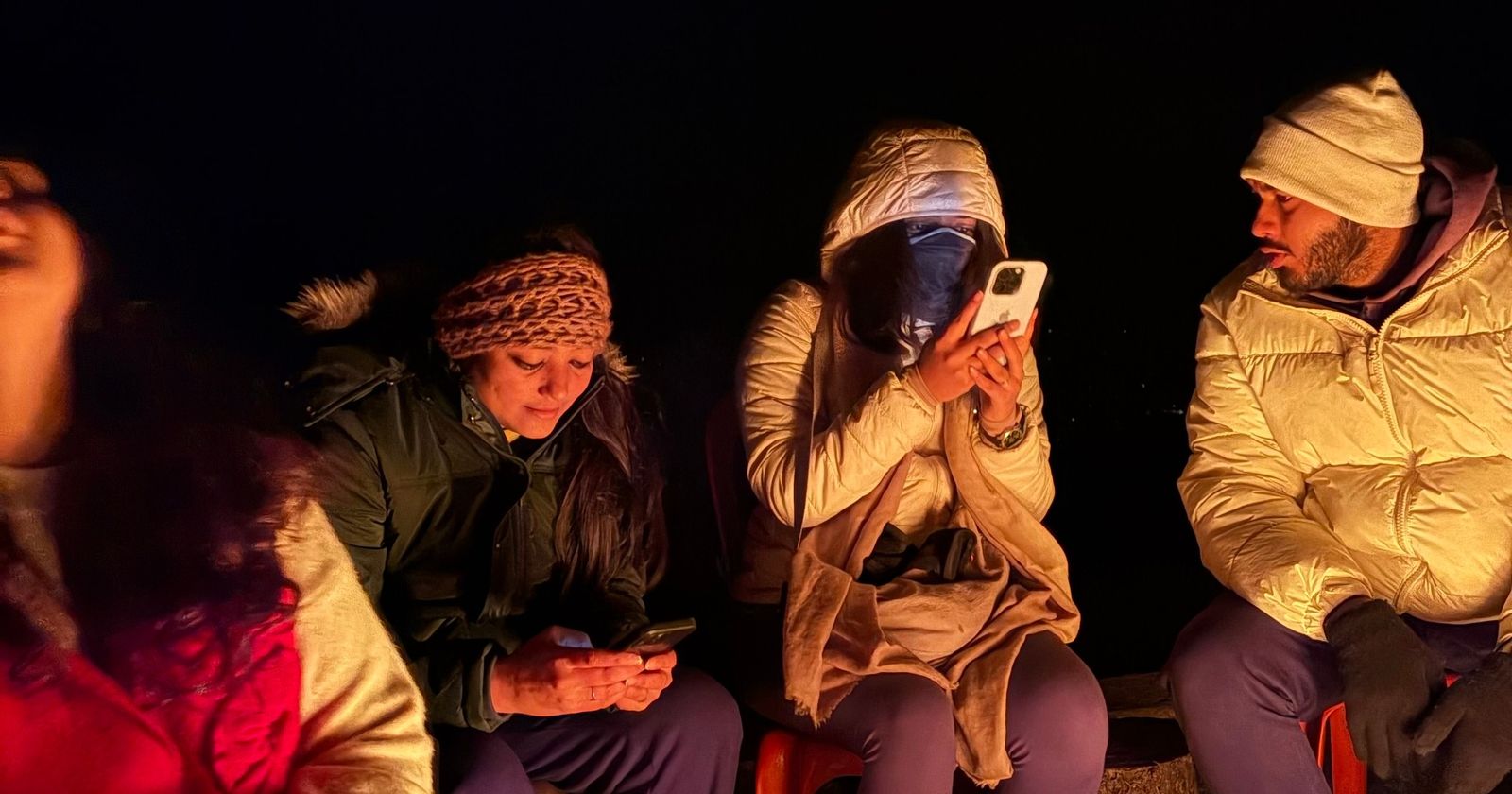 Friends in winter jackets sitting around a bonfire at night in the mountains
